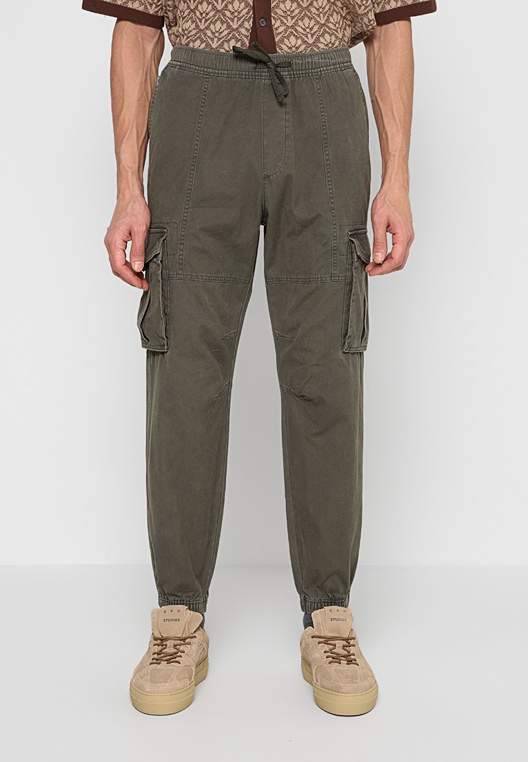 Mann trägt olivgrüne Cargo-Jogginghosen mit elastischem Bund und Bündchen, kombiniert mit beigen Sneakers und einem gemusterten Hemd.