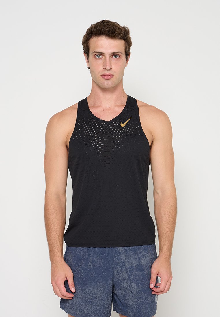 Junger Mann trägt ein schwarzes ärmelloses sportliches Tanktop mit goldenem Nike-Logo und blaue Sportshorts und steht vor einem einfarbigen Hintergrund.
