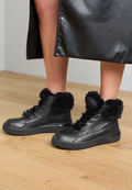 Schwarze High-Top-Sneaker mit Kunstpelzkragen, Obermaterial aus Leder, Gummisohlen und Schnürdesign, auf einem Holzfußboden.