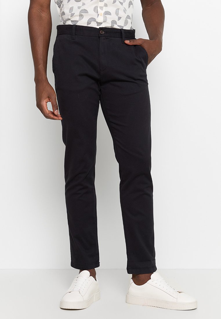 Schwarze Baumwollhose im Slim-Fit-Stil mit Knopfverschluss, seitlichen Taschen und glatter Textur, kombiniert mit weißen Sneakers.