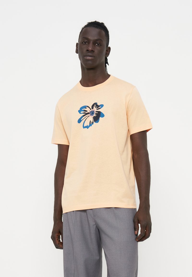 Mann trägt ein hellpfirsichfarbenes T-Shirt mit einem blauen und schwarzen abstrakten Blumendesign, kombiniert mit grauen Hosen, stehend vor einem einfarbigen Hintergrund.