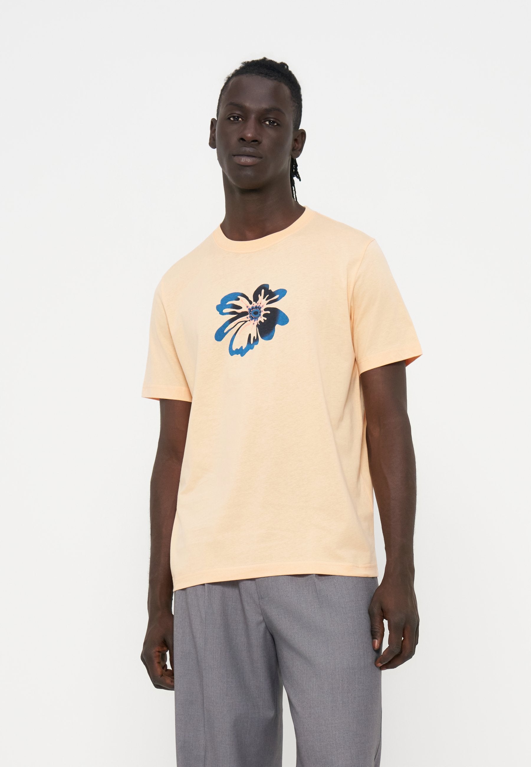 Mann trägt ein hellpfirsichfarbenes T-Shirt mit einem blauen und schwarzen abstrakten Blumendesign, kombiniert mit grauen Hosen, stehend vor einem einfarbigen Hintergrund.
