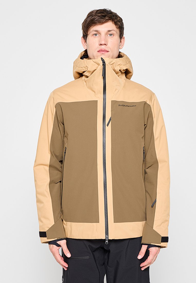 Junger Mann trägt eine beige-olivgrüne Outdoorjacke mit Kapuze und Frontreißverschluss und schwarze Hose, steht vor einem schlichten Hintergrund.