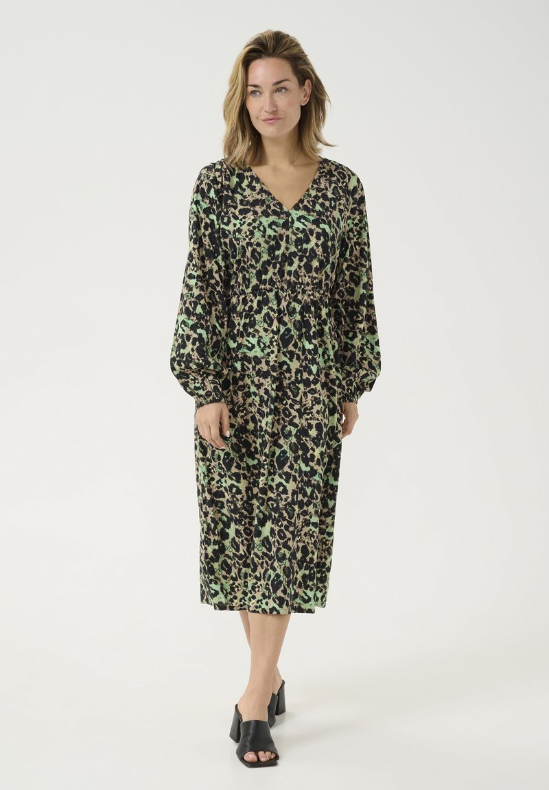 Leopardenmuster-Kleid in Grün und Schwarz, mit V-Ausschnitt, langen Ärmeln und elastischem Bund. Kombiniert mit schwarzen, offenen Absatzschuhen.
