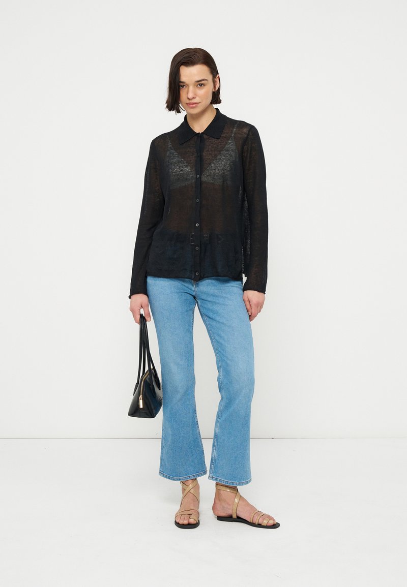 Frau mit kurzen dunklen Haaren, die ein transparentes schwarzes Hemd mit Knöpfen, blaue kurze Jeans, goldene Sandalen trägt und eine schwarze Handtasche hält.